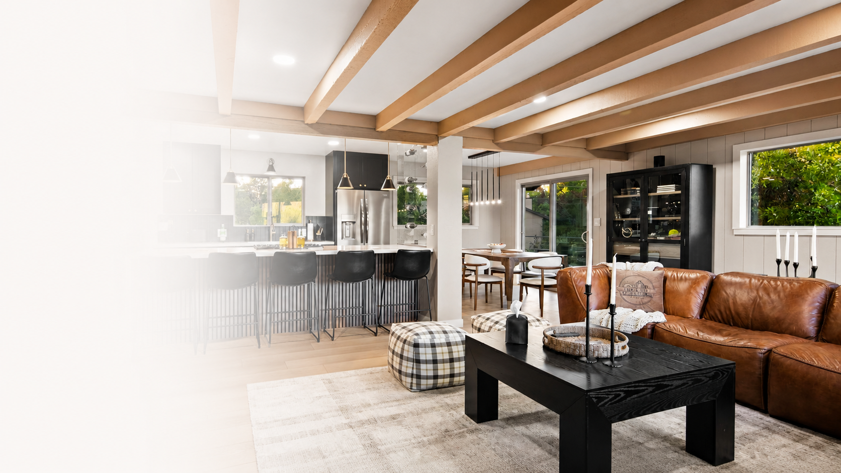 An open-plan vacation rental interior: a leather sectional and black coffee table in the foreground, with a kitchen island, bar stools and a dining area beyond, under exposed wood ceiling beams