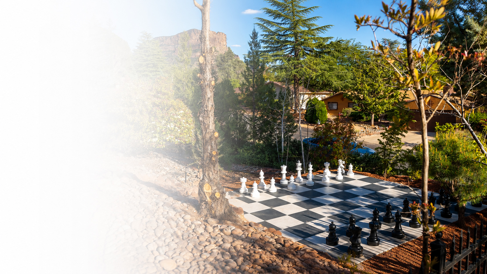A giant outdoor chess set on a black-and-white patio board at a vacation rental, surrounded by trees, neighboring houses and red-rock cliffs in the sunshine